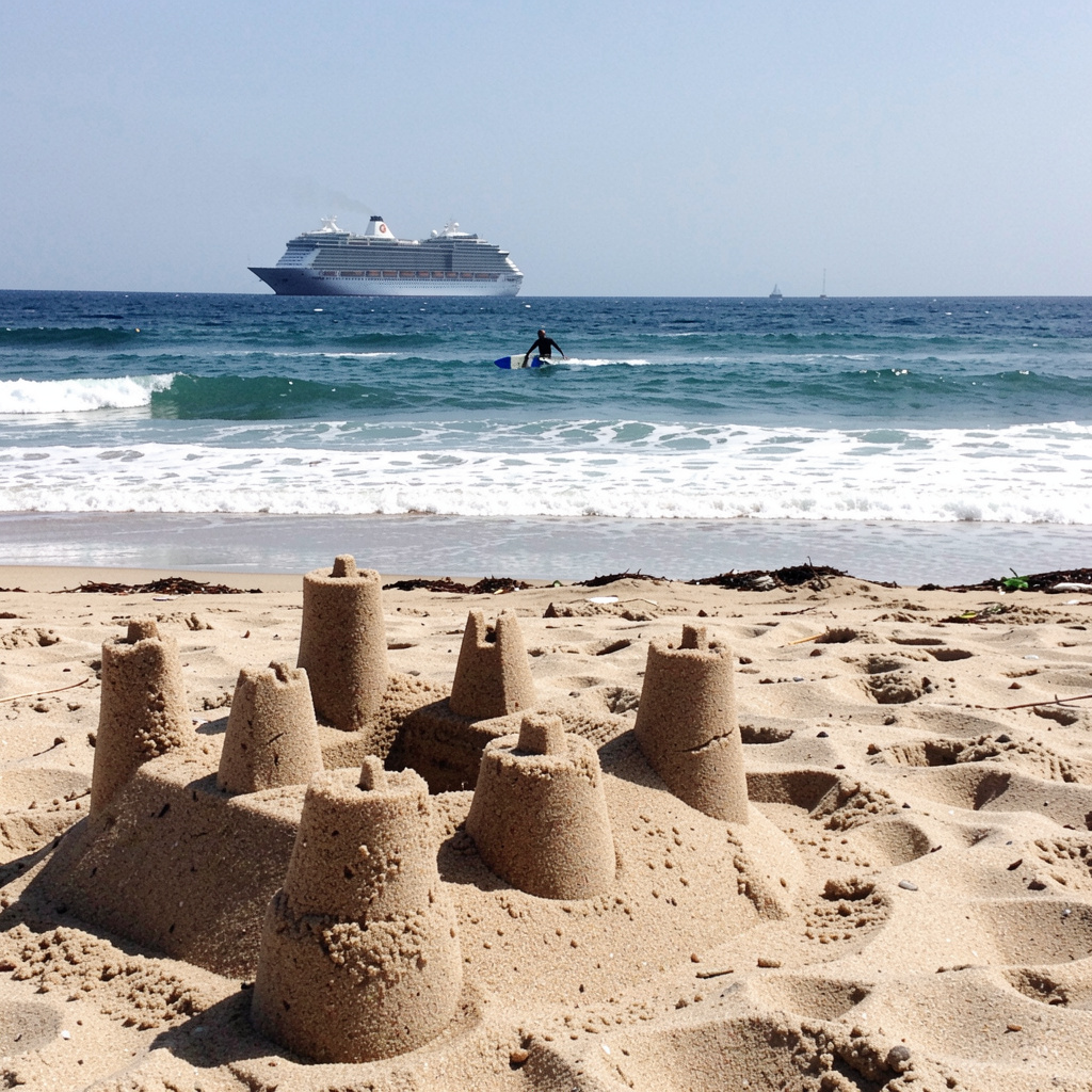 A beach scene with a sandcastle in the foreground, a surfer 